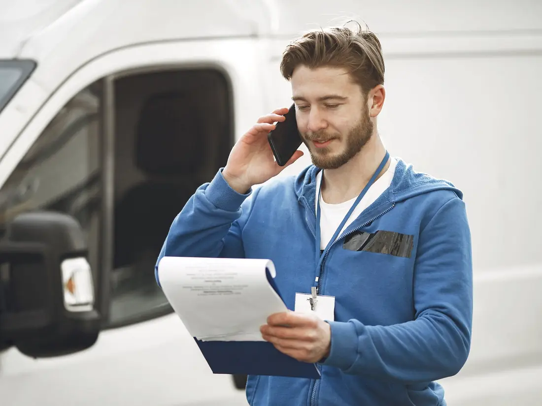 A tradesperson standing at a van, taking a phone call while holding a job sheet.