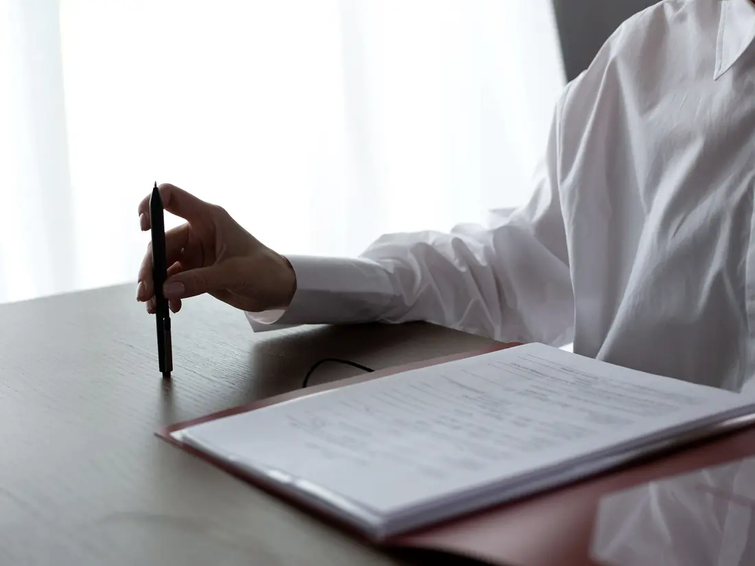 A hand resting a pen on a printed document on a desk in window light.