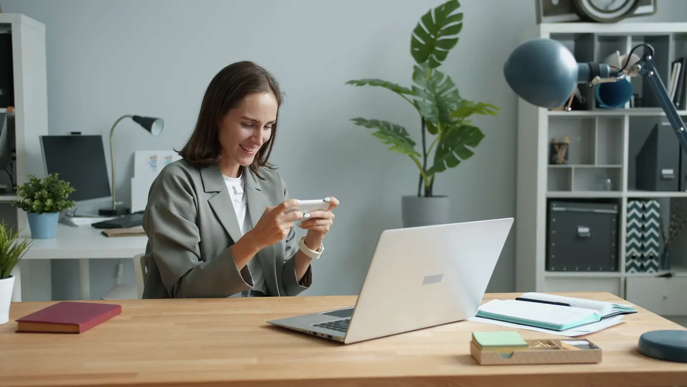 Someone at a desk reading a message on their phone, with a laptop open in front of them.